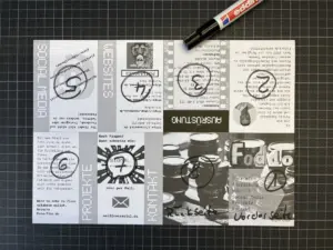 Ein schwarz-weiß gedrucktes Flugblatt auf einer gerasterten Fläche, markiert mit Kreisen und Zahlen in schwarzem Marker; darüber liegt ein Stift.