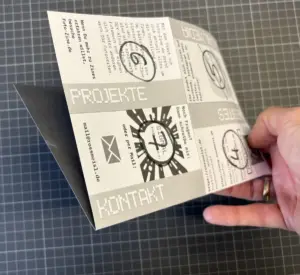 Eine Hand hält eine grau bedruckte Broschüre mit den Aufschriften "PROJEKTE" und "KONTAKT" auf einer gitterartigen Oberfläche.