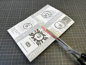 Eine Hand schneidet mit einer Schere einen grauen Falzflyer mit Text und Grafiken auf einer schwarzen Gitterfläche aus.