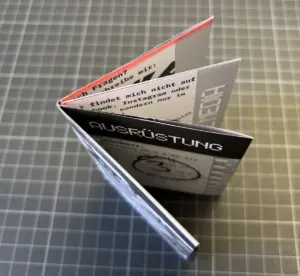 Ein kleines gefaltetes Zine mit deutschem Text, von oben gesehen auf einer grauen, gitterartigen Oberfläche.
